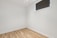 A small, empty room with light wooden flooring and plain white walls. A single high window with a dark blind lets in minimal light, creating a minimalist and sterile appearance.