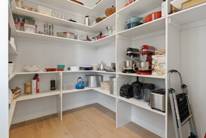 A pantry with white shelving contains various kitchen appliances and storage items. Mixing bowls, baking trays, and small kitchen gadgets are neatly organized. On the shelves are mixer machines, toasters, and a variety of pots. Additionally, there are containers for dry goods, a fire extinguisher, and a folding step ladder.