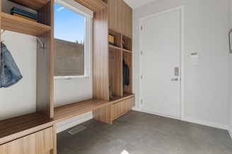 A clean and modern entryway with wooden built-in storage units, including open cubbies, shelves, and a bench. There is a white door with a digital lock on the right side, and a window with a view of the outdoors on the left. The floor is tiled with gray tiles, and a few items of clothing are hung in the cubbies.