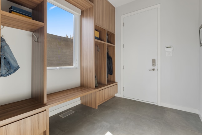 A clean and modern entryway with wooden built-in storage units, including open cubbies, shelves, and a bench. There is a white door with a digital lock on the right side, and a window with a view of the outdoors on the left. The floor is tiled with gray tiles, and a few items of clothing are hung in the cubbies.