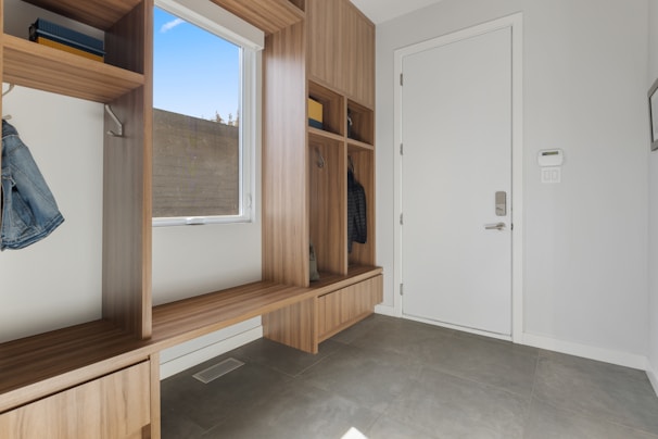 A clean and modern entryway with wooden built-in storage units, including open cubbies, shelves, and a bench. There is a white door with a digital lock on the right side, and a window with a view of the outdoors on the left. The floor is tiled with gray tiles, and a few items of clothing are hung in the cubbies.