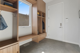 A clean and modern entryway with wooden built-in storage units, including open cubbies, shelves, and a bench. There is a white door with a digital lock on the right side, and a window with a view of the outdoors on the left. The floor is tiled with gray tiles, and a few items of clothing are hung in the cubbies.