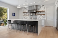 A modern kitchen with a large white island and black bar stools. The backsplash features a marble design, and stainless steel appliances are prominent. Open shelves display kitchen items, and there's a piece of artwork on the wall. Natural light streams in from the large window, revealing greenery outside.