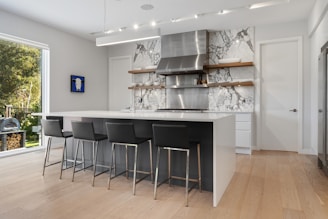 A modern kitchen with a large white island and black bar stools. The backsplash features a marble design, and stainless steel appliances are prominent. Open shelves display kitchen items, and there's a piece of artwork on the wall. Natural light streams in from the large window, revealing greenery outside.