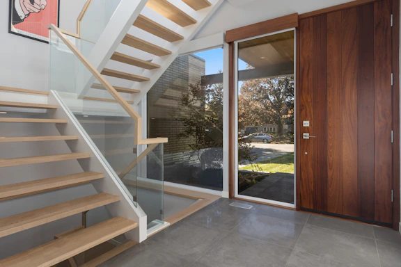A modern interior entryway with a wooden staircase featuring glass panels and light wood steps. The area includes a wooden door with a large glass window adjacent to it, providing a clear view of the outside. The floor is tiled with large, gray tiles, and there is a minimalist art piece on the wall.