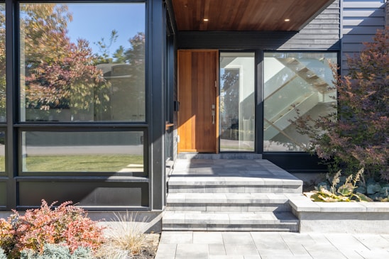 The image features the entrance of a modern house with large glass windows and a wooden front door. The entryway has gray stone steps leading up to the door, and the area is bordered by a small garden with various plants and shrubs. The exterior walls are a combination of black siding and wood paneling, creating a sleek and contemporary look.