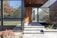 The image features the entrance of a modern house with large glass windows and a wooden front door. The entryway has gray stone steps leading up to the door, and the area is bordered by a small garden with various plants and shrubs. The exterior walls are a combination of black siding and wood paneling, creating a sleek and contemporary look.