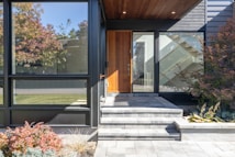 The image features the entrance of a modern house with large glass windows and a wooden front door. The entryway has gray stone steps leading up to the door, and the area is bordered by a small garden with various plants and shrubs. The exterior walls are a combination of black siding and wood paneling, creating a sleek and contemporary look.