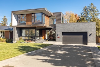 A modern two-story house with a sleek architectural design, featuring large windows and a combination of wood and dark brick exterior. The house has a spacious driveway and a manicured lawn. Trees with autumn foliage are visible in the background, adding a touch of nature to the setting.