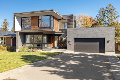A modern two-story house with a sleek architectural design, featuring large windows and a combination of wood and dark brick exterior. The house has a spacious driveway and a manicured lawn. Trees with autumn foliage are visible in the background, adding a touch of nature to the setting.