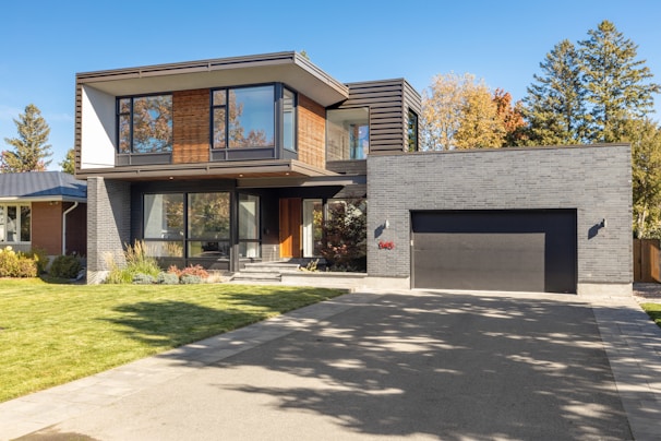 A modern two-story house with a sleek architectural design, featuring large windows and a combination of wood and dark brick exterior. The house has a spacious driveway and a manicured lawn. Trees with autumn foliage are visible in the background, adding a touch of nature to the setting.