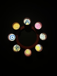 A circular arrangement of eight small round artworks is set against a dark background. Each piece features unique abstract designs with colorful patterns, including geometric shapes, heart motifs, and swirling lines.