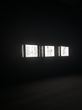 Three illuminated screens are set against a dark background, each displaying hand-drawn illustrations of various objects or scenes. The images on the screens appear to be simplistic line drawings, possibly resembling abstract art or sketches.