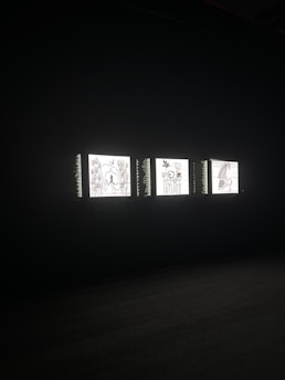 Three illuminated screens are set against a dark background, each displaying hand-drawn illustrations of various objects or scenes. The images on the screens appear to be simplistic line drawings, possibly resembling abstract art or sketches.