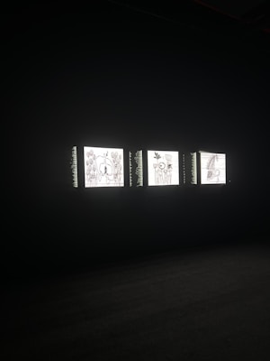 Three illuminated screens are set against a dark background, each displaying hand-drawn illustrations of various objects or scenes. The images on the screens appear to be simplistic line drawings, possibly resembling abstract art or sketches.