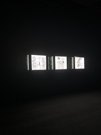 Three illuminated screens are set against a dark background, each displaying hand-drawn illustrations of various objects or scenes. The images on the screens appear to be simplistic line drawings, possibly resembling abstract art or sketches.