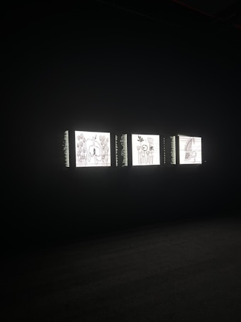 Three illuminated screens are set against a dark background, each displaying hand-drawn illustrations of various objects or scenes. The images on the screens appear to be simplistic line drawings, possibly resembling abstract art or sketches.