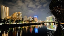 A nighttime cityscape with illuminated buildings reflecting on a calm river. The sky is cloudy, and city lights create vibrant reflections in the water. Trees line the riverbank, and there is a modern urban vibe with well-lit high-rises.