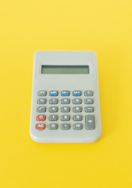 A simple electronic calculator with a gray body and standard number and function keys. It is set against a bright yellow background, highlighting the calculator as the central focus of the image.