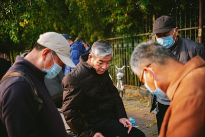 A group of elderly men are gathered outdoors, wearing jackets and face masks, possibly engaged in a conversation or playing a game. They appear to be in a park with trees and a fence in the background. The lighting suggests a sunny day.