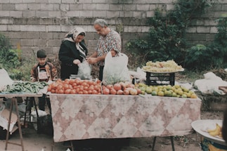 A family is gathered around a market stall filled with fresh produce, including tomatoes, green peppers, limes, and grapes. The father is bagging some items for a customer, while the mother and son look on. The scene is set against a rough stone wall with foliage growing in the background.