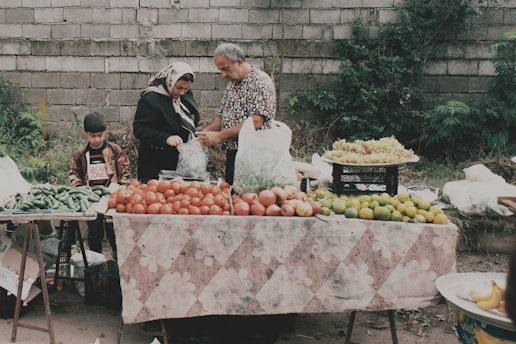 A family is gathered around a market stall filled with fresh produce, including tomatoes, green peppers, limes, and grapes. The father is bagging some items for a customer, while the mother and son look on. The scene is set against a rough stone wall with foliage growing in the background.