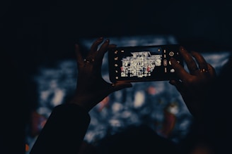 A person holding a smartphone, capturing a photograph of a structure displayed on its screen. The scene is set in low light, focusing on the illuminated phone screen amidst a dark background.