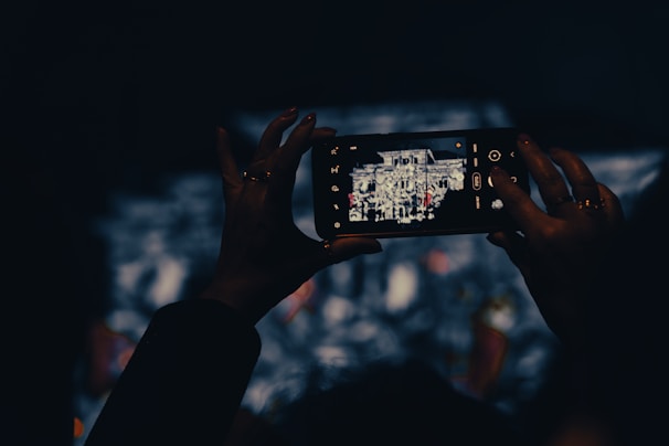 A person holding a smartphone, capturing a photograph of a structure displayed on its screen. The scene is set in low light, focusing on the illuminated phone screen amidst a dark background.