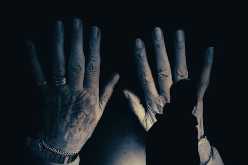 Two large, weathered hands are prominently featured, positioned against a dark background. The hands, adorned with rings and a watch, appear aged and textured, suggesting a history of hard work. In the foreground, a silhouette of a person is visible, seemingly gazing at the hands.