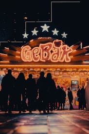 A brightly lit carnival or fairground stall with the word 'GEBÄK' prominently displayed in neon lights. Silhouettes of people are gathered around the stall, giving a sense of activity and interest. The background includes stars illuminated above the stall.