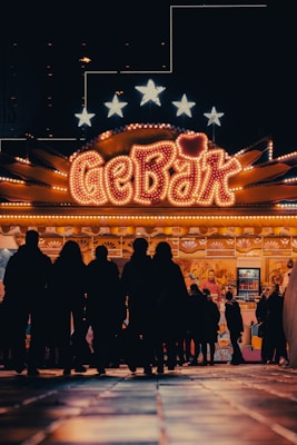 A brightly lit carnival or fairground stall with the word 'GEB&Auml;K' prominently displayed in neon lights. Silhouettes of people are gathered around the stall, giving a sense of activity and interest. The background includes stars illuminated above the stall.