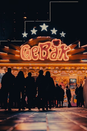 A brightly lit carnival or fairground stall with the word 'GEB&Auml;K' prominently displayed in neon lights. Silhouettes of people are gathered around the stall, giving a sense of activity and interest. The background includes stars illuminated above the stall.