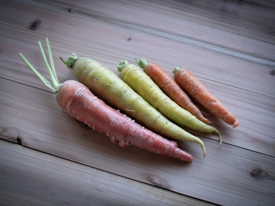 A group of six carrots of varying colors, including pink, greenish-yellow, and orange, are arranged on a light wooden surface. The carrots are different sizes and display natural textures and imperfections.