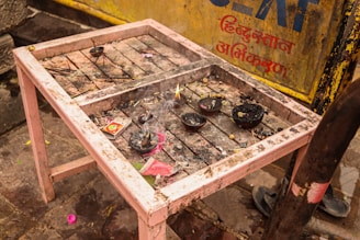 A small, weathered table holds several burnt offerings, including incense sticks and charred bowls. A burnt matchbox and remnants of ash are scattered across the dusty surface. In the background, there is a yellow metal panel with Hindi script.