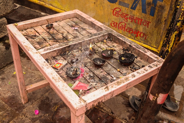 A small, weathered table holds several burnt offerings, including incense sticks and charred bowls. A burnt matchbox and remnants of ash are scattered across the dusty surface. In the background, there is a yellow metal panel with Hindi script.