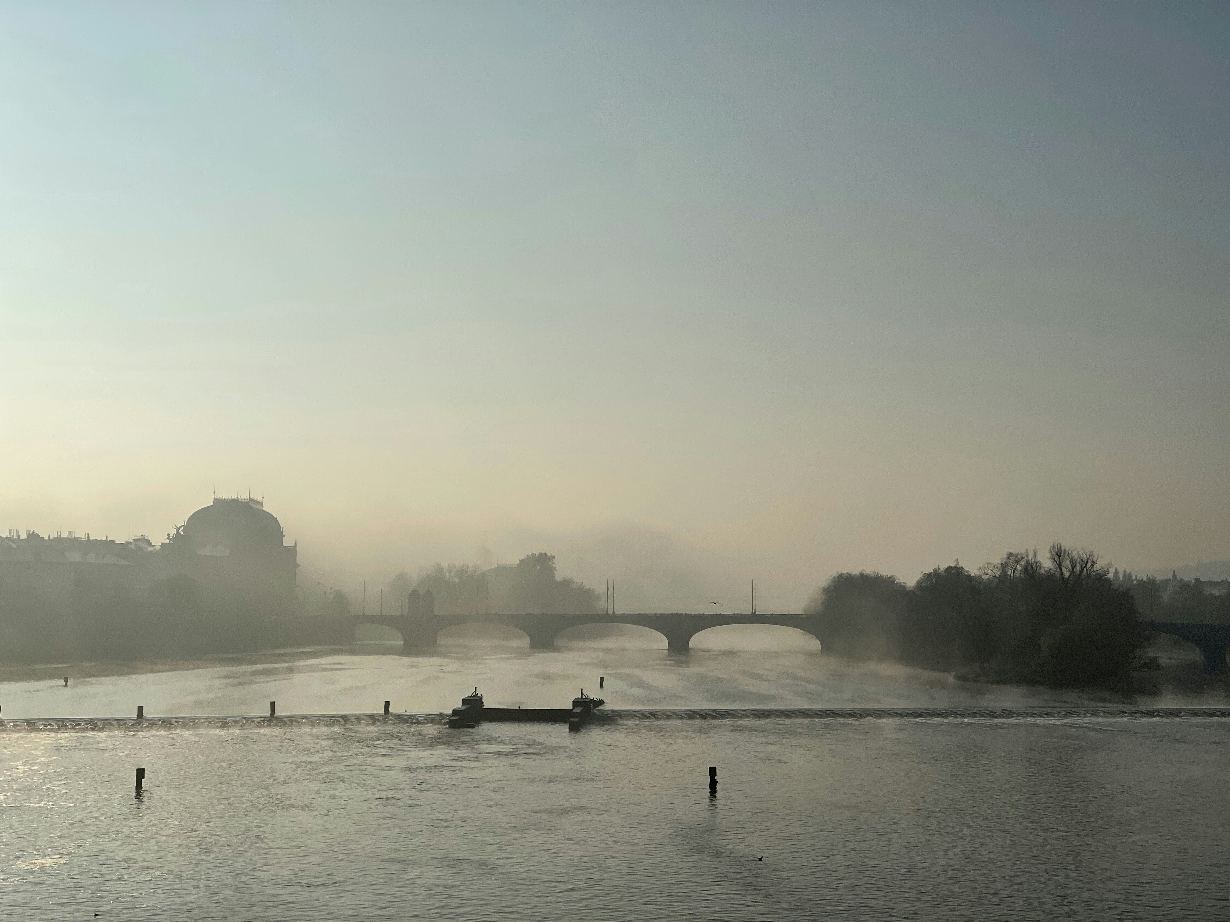 A misty November morning on Prague's Charles Bridge, with the Vltava River dominating the view. Looking south, the historic Legion Bridge (Most Legií) and the silhouette of the National Theatre stand out in the distance.