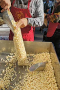 A person is filling a plastic bag with popcorn from a large metal tray using a metallic scoop. The individual is wearing a red apron and a grey sweatshirt. There are other people visible in the background, indicating a market or event setting.
