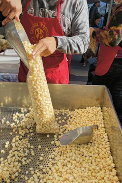 A person is filling a plastic bag with popcorn from a large metal tray using a metallic scoop. The individual is wearing a red apron and a grey sweatshirt. There are other people visible in the background, indicating a market or event setting.