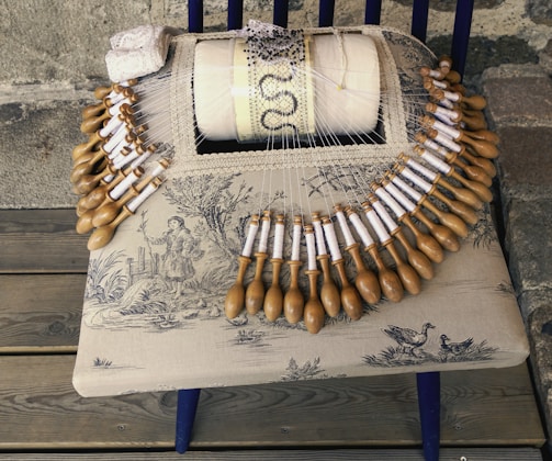 A traditional bobbin lace-making setup on a wooden chair, featuring a pillow with intricate lacework and numerous wooden bobbins arranged in a semicircle. The chair's seat is upholstered with fabric depicting a pastoral scene with a figure and birds. A small piece of completed lace and some beads are also visible.