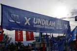 A large blue banner with the words 'Undilah Barisan Nasional' and a logo featuring a scale, possibly indicating a political campaign. Surrounding are other flags and banners, mainly red and blue, set against a partly cloudy sky and some visible trees and buildings in the background.