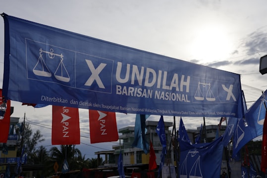 A large blue banner with the words 'Undilah Barisan Nasional' and a logo featuring a scale, possibly indicating a political campaign. Surrounding are other flags and banners, mainly red and blue, set against a partly cloudy sky and some visible trees and buildings in the background.