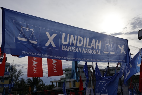 A large blue banner with the words 'Undilah Barisan Nasional' and a logo featuring a scale, possibly indicating a political campaign. Surrounding are other flags and banners, mainly red and blue, set against a partly cloudy sky and some visible trees and buildings in the background.