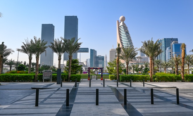 A modern cityscape featuring tall skyscrapers and distinctive architecture, with palm trees and green foliage lining a paved urban walkway. The sky is clear and blue, contributing to the vibrant urban atmosphere.