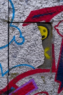 A textured concrete surface features vibrant graffiti art with various abstract shapes and colors. The image includes a prominent red shape resembling a cap, a yellow circular form suggesting an eye, and swirling blue and red lines.