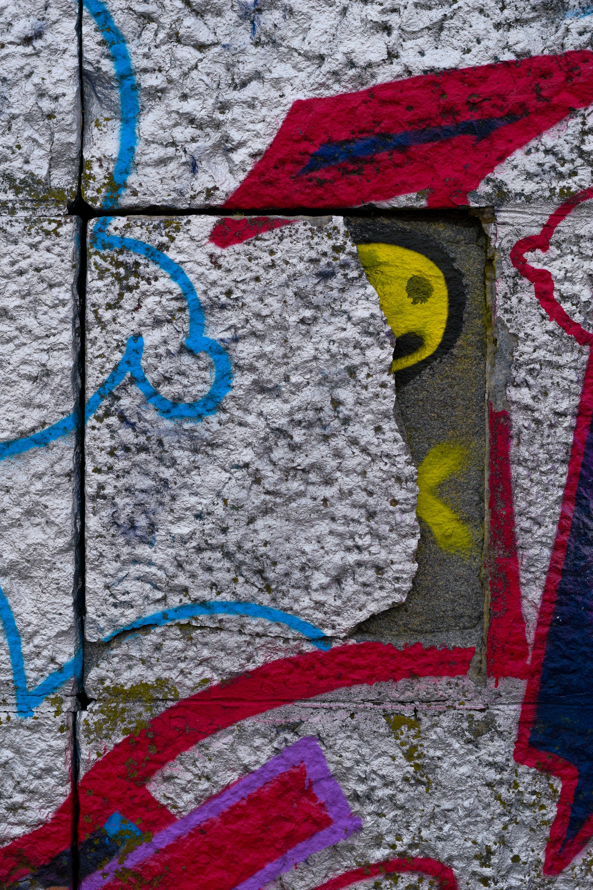 A textured concrete surface features vibrant graffiti art with various abstract shapes and colors. The image includes a prominent red shape resembling a cap, a yellow circular form suggesting an eye, and swirling blue and red lines.