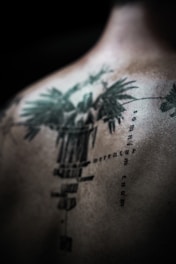 An abstract, detailed tattoo is depicted on a person's back, featuring intricate designs and text in a vertical alignment. The tattoo has a dark green to black color scheme, with sharp, defined lines and some blurred areas, creating a mysterious and artistic effect.