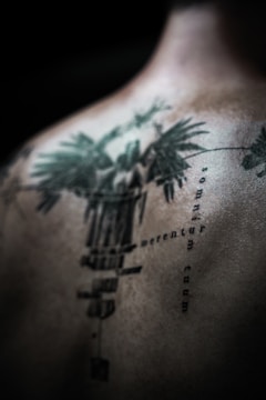An abstract, detailed tattoo is depicted on a person's back, featuring intricate designs and text in a vertical alignment. The tattoo has a dark green to black color scheme, with sharp, defined lines and some blurred areas, creating a mysterious and artistic effect.