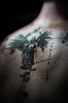 An abstract, detailed tattoo is depicted on a person's back, featuring intricate designs and text in a vertical alignment. The tattoo has a dark green to black color scheme, with sharp, defined lines and some blurred areas, creating a mysterious and artistic effect.