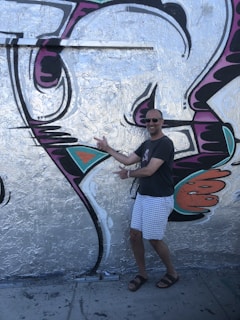 A man in a casual outfit, including a dark t-shirt, light checkered shorts, and sandals, is posing enthusiastically in front of a large piece of graffiti. The artwork is vibrant with silver, black, purple, orange, and teal colors forming abstract shapes on a wall.