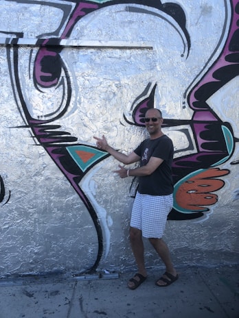 A man in a casual outfit, including a dark t-shirt, light checkered shorts, and sandals, is posing enthusiastically in front of a large piece of graffiti. The artwork is vibrant with silver, black, purple, orange, and teal colors forming abstract shapes on a wall.